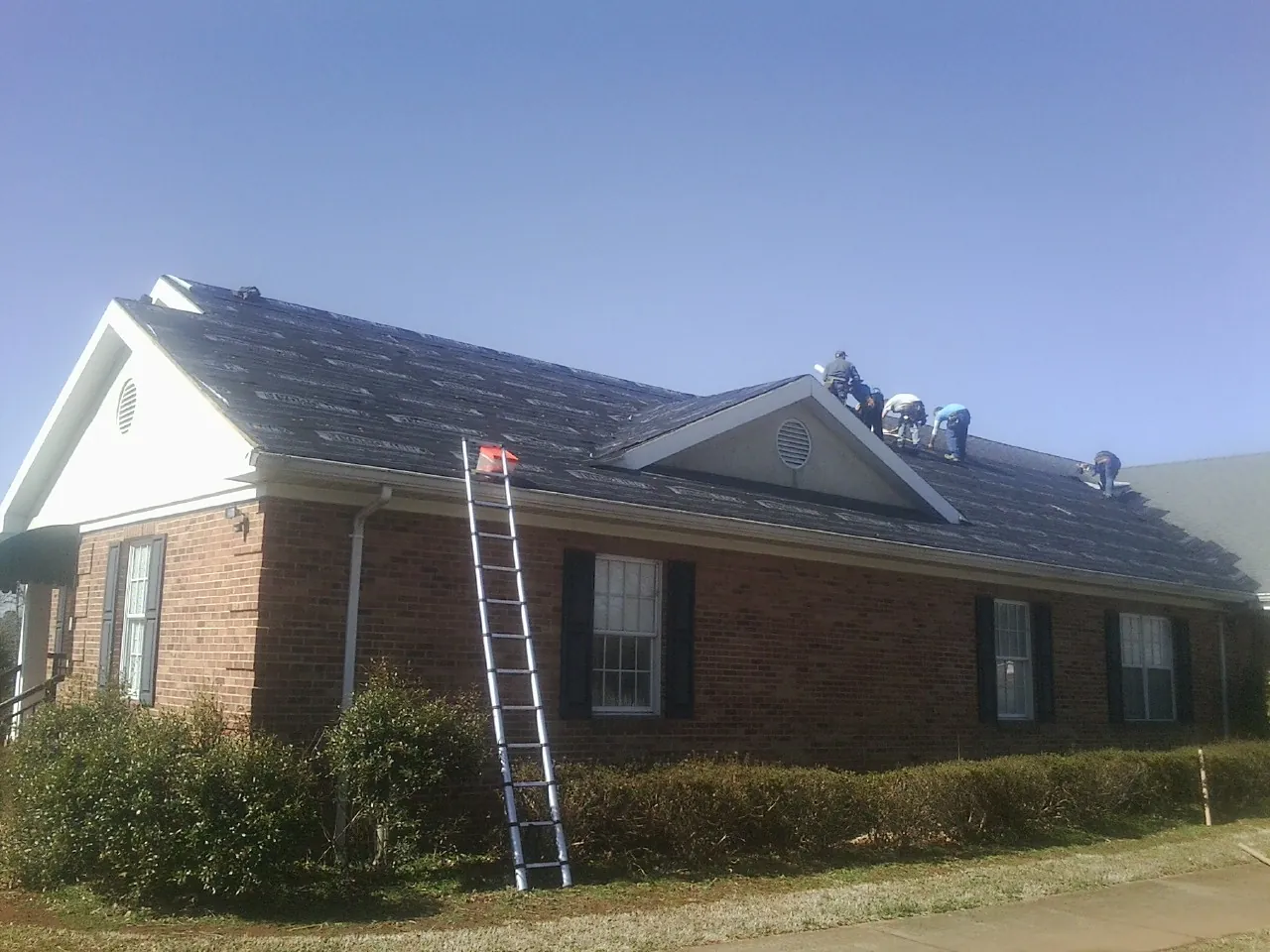 Crew tearing shingles off a brick ranch roof with a ladder set — Emergency Roof Repair work by our crew in Davisburg