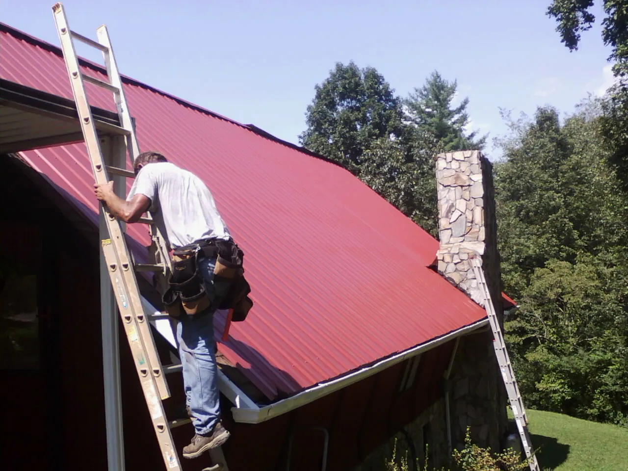 Roofer on a ladder beside a red metal roof and stone chimney — Soffit & Fascia Repair by Roofing Brothers in Davisburg